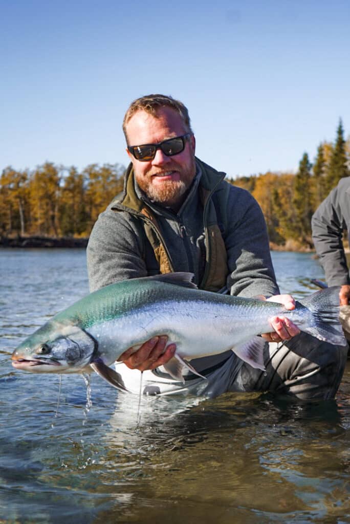 A man wearing sunglasses and outdoor clothes stands in shallow water, smiling and holding a large silver fish. Trees with autumn foliage and a clear blue sky are visible behind him—ideal scenery for those seeking hunting property or land for sale.