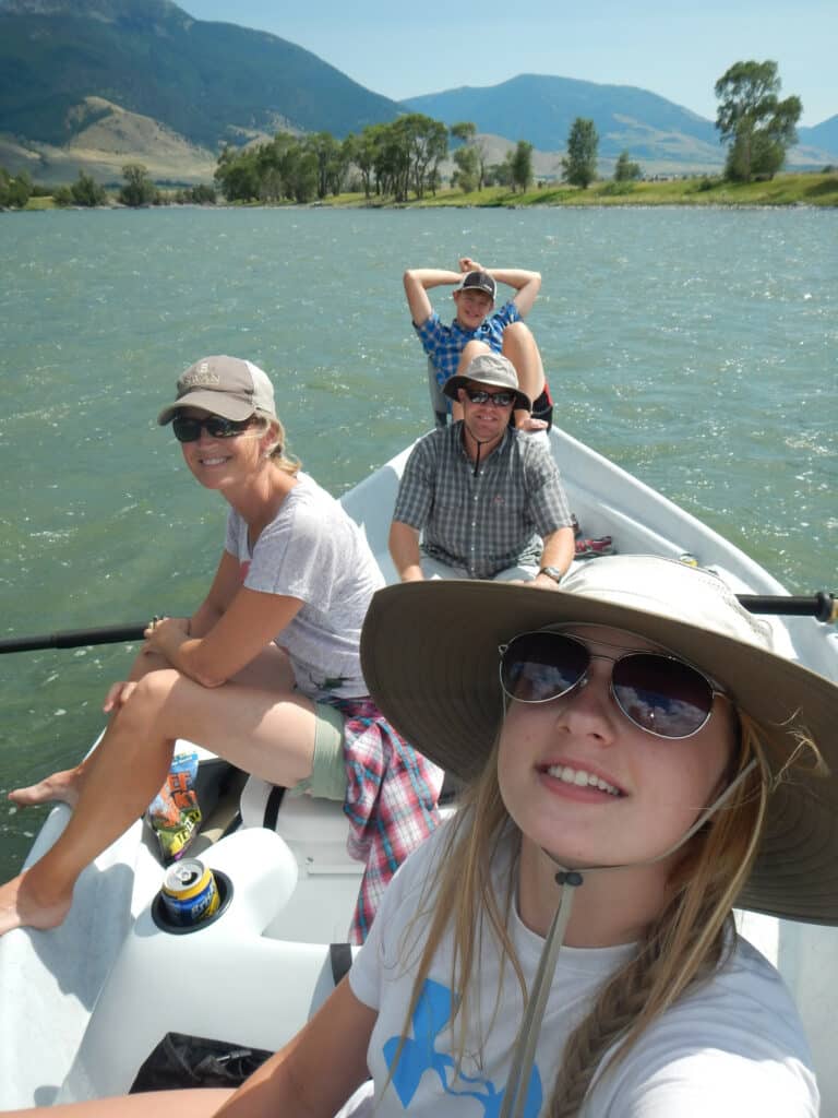 Four people enjoy a sunny day on a lake, surrounded by mountains and trees on recreational land. Wearing hats and sunglasses, they smile and relax as they pose for a group selfie on the water.