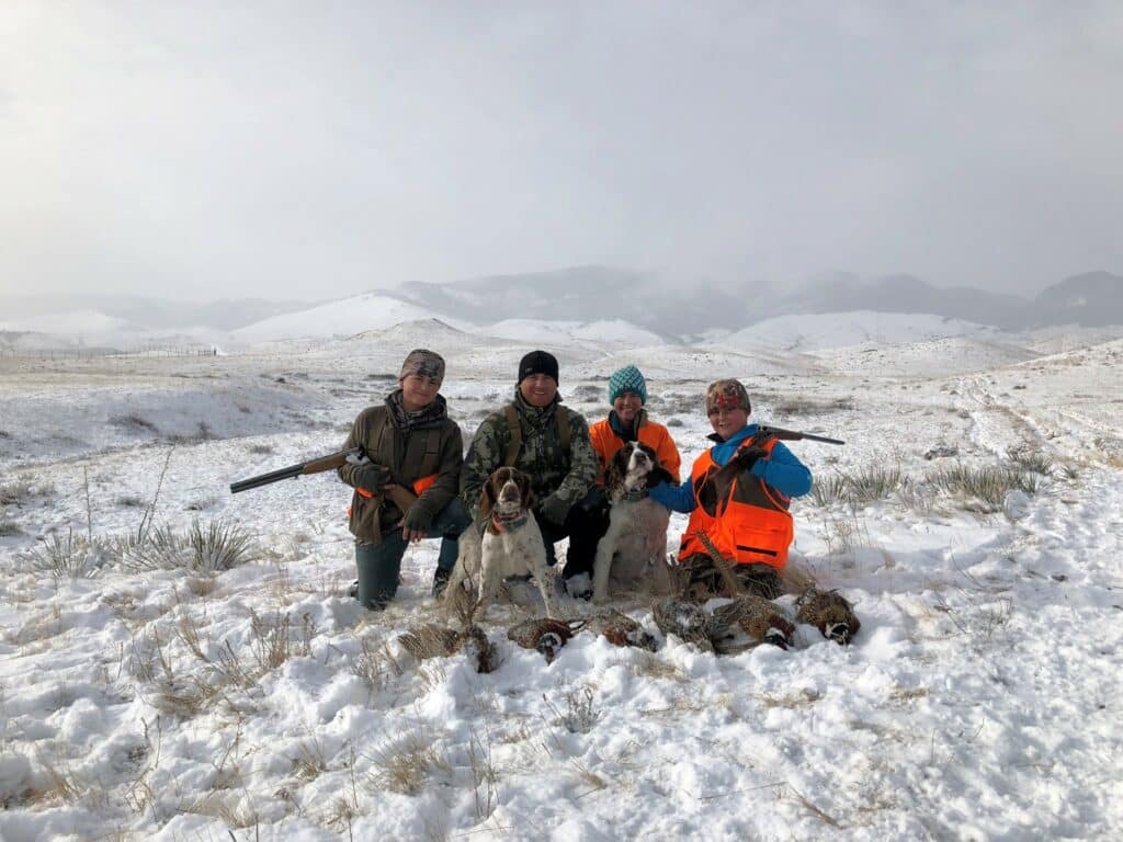 Four people in winter gear kneel in a snowy field on recreational land with two dogs, holding rifles and displaying several birds. Snow-covered hills and mountains are visible in the background under a cloudy sky.