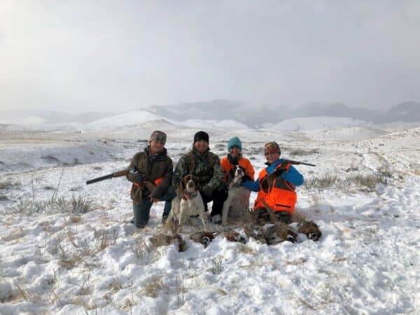 Four people in winter gear kneel in a snowy field on recreational land with two dogs, holding rifles and displaying several birds. Snow-covered hills and mountains are visible in the background under a cloudy sky.