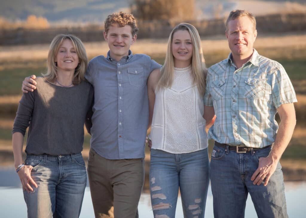 A family of four stands outdoors in casual clothes, smiling and posing together on a grassy field with a pond in the background—perfect for those seeking hunting property or land for sale.