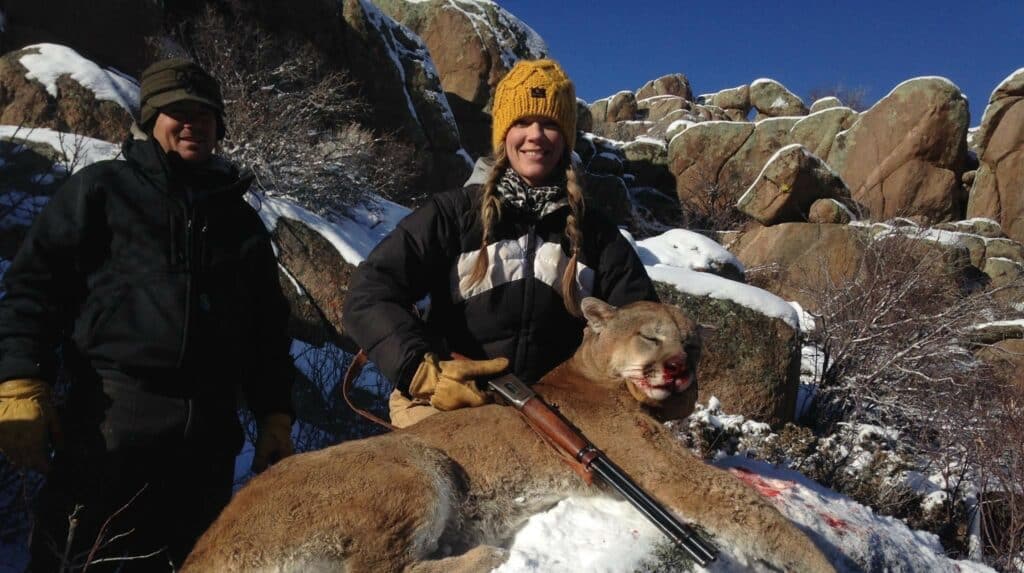 Two people in winter clothing pose outdoors on a snowy, rocky cattle ranch with a large, deceased mountain lion in the foreground; one holds a rifle and smiles at the camera, showcasing the hunting property.