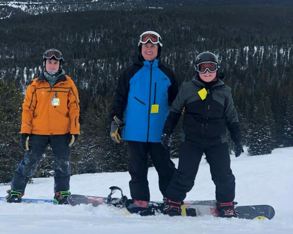 Three people wearing winter gear and helmets stand on a snowy slope with snowboards at their feet, overlooking a forested mountain landscape perfect for hunting property or land for sale.