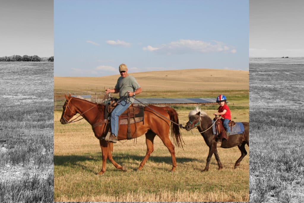 An adult and a child ride horses in a grassy field on a cattle ranch. The adult is on a brown horse, sunglasses and cap on, while the child follows behind with a helmet. Rolling hills and blue sky complete the scenic backdrop.