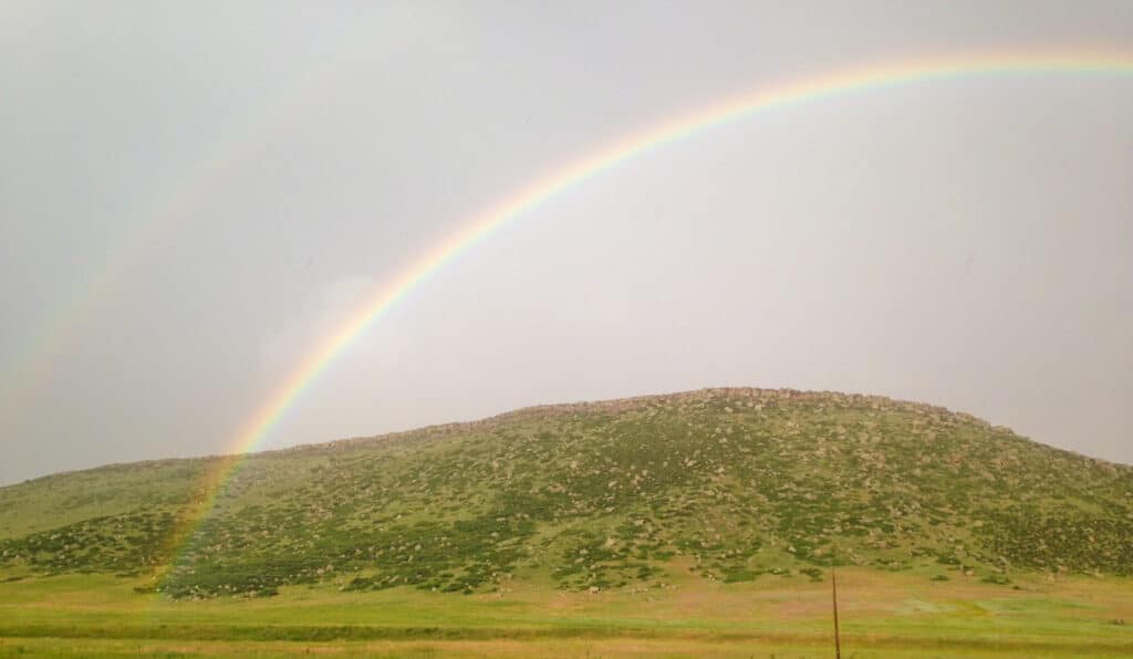 A vibrant rainbow arcs across a cloudy sky above a green, rocky hill in an open grassy landscape—perfect recreational land. The faint outline of a second rainbow is visible to the left.