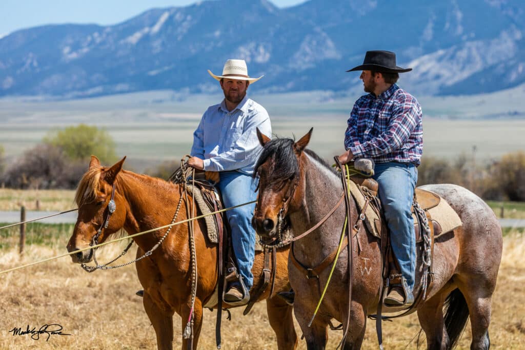 Two men wearing hats and plaid shirts ride horses across recreational land in an open field with mountains behind them on a sunny day. They appear to be talking to each other.