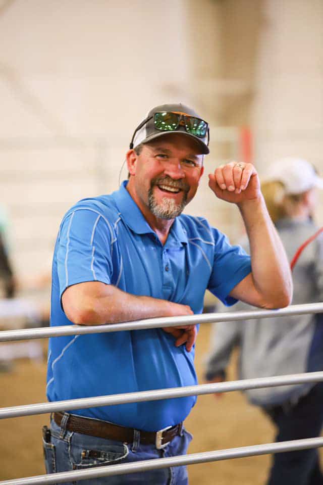 A man in a blue polo shirt and sunglasses smiles while leaning on a metal fence indoors, with people and blurred background behind him—capturing the relaxed feel of a cattle ranch or hunting property.
