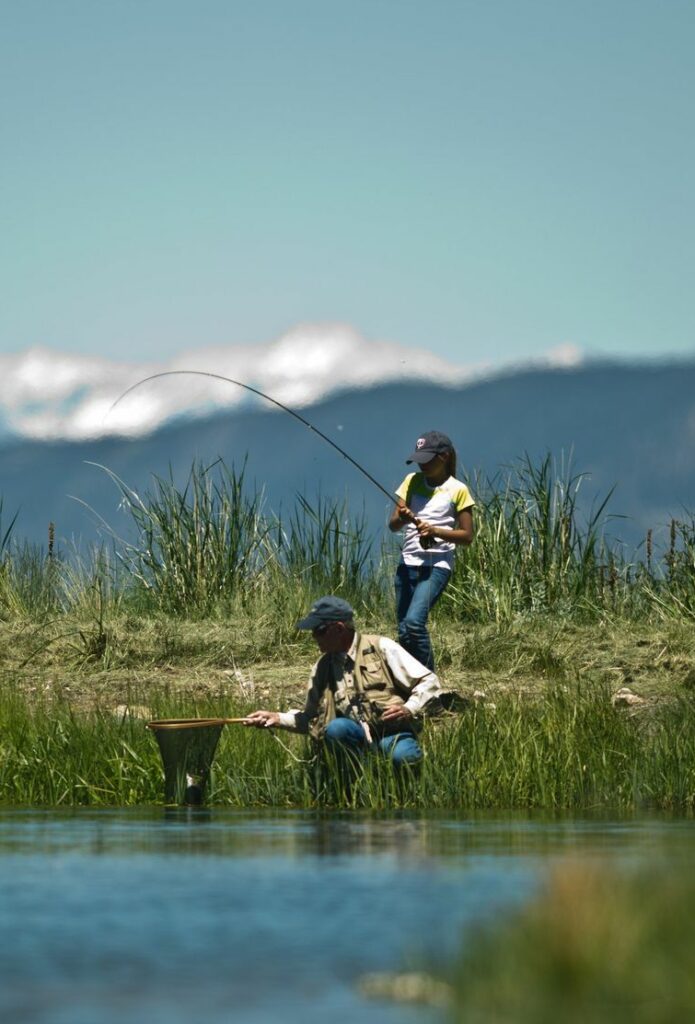 Two people fish by a lake surrounded by tall grass on recreational land. One stands with a fishing rod, while the other kneels by the water with a net. Snow-capped mountains and blue sky form the scenic backdrop.