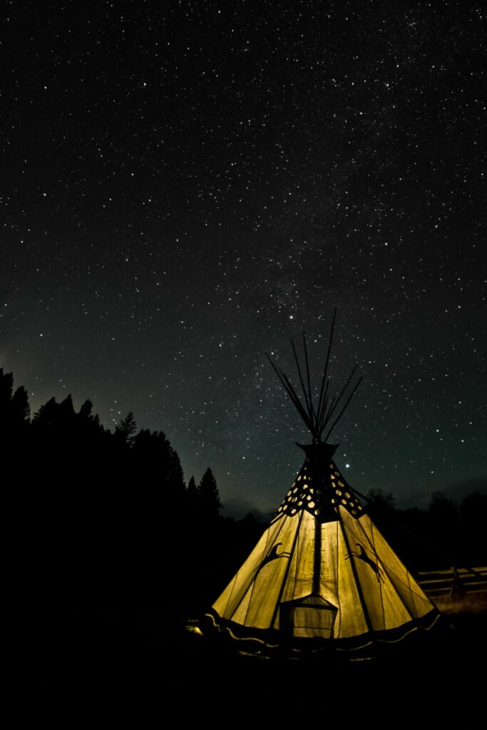 A warmly lit tipi stands beneath a clear, star-filled night sky on a tranquil hunting property, surrounded by silhouettes of trees and forest.