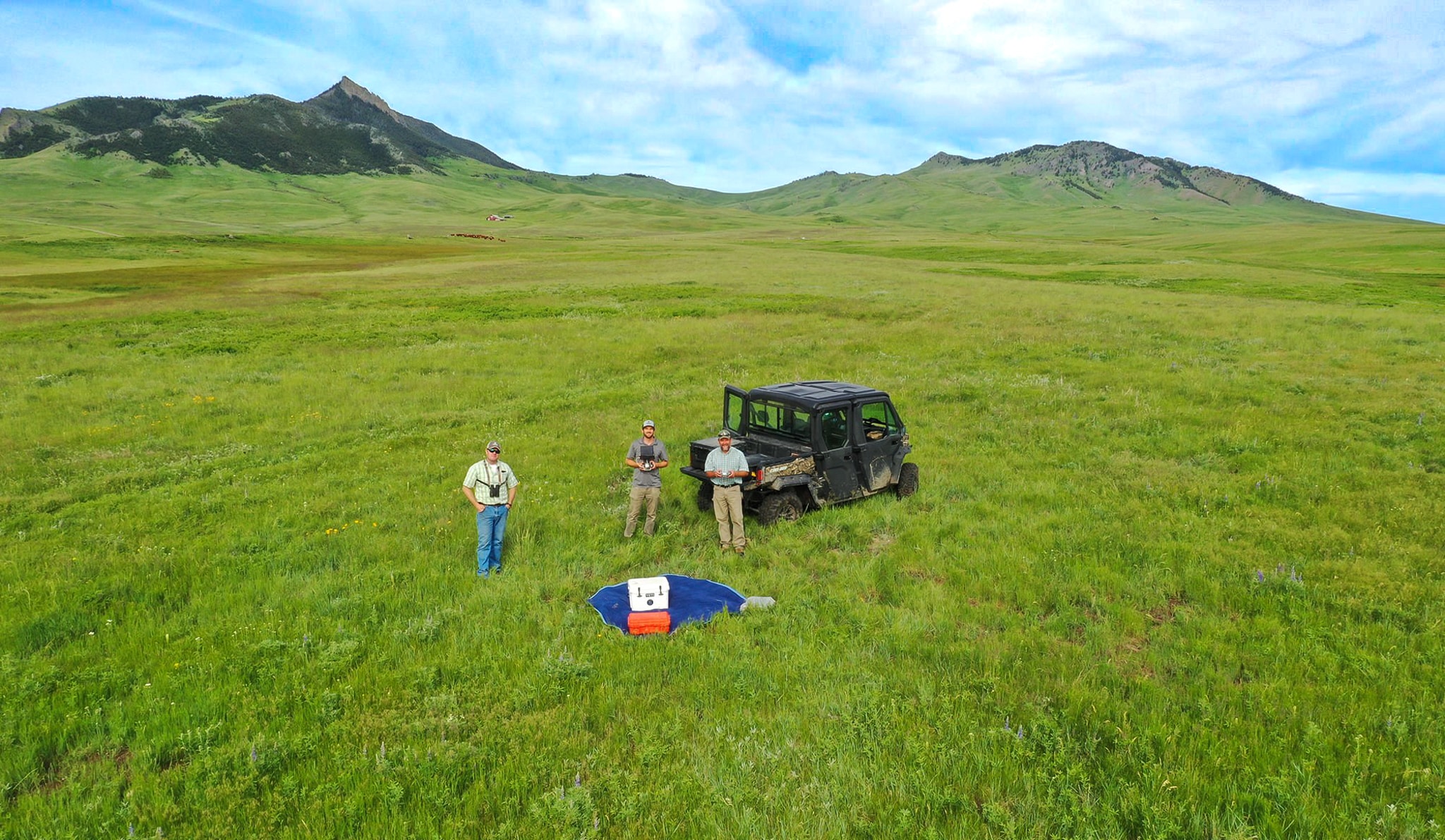 Three people stand in a grassy field near an off-road vehicle, with a blue and white parachute and equipment on the ground—perfect recreational land or hunting property surrounded by rolling green hills and mountains under a blue sky.