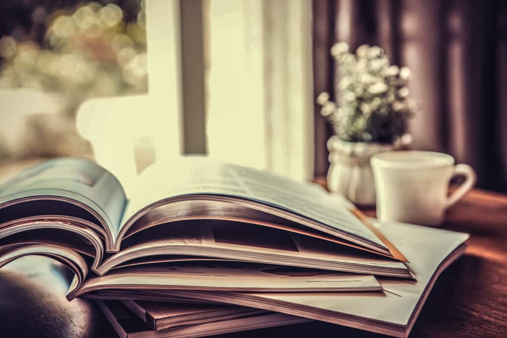 A stack of open books on a wooden table beside a white cup and a small vase with white flowers, sunlight streaming in through the window, evokes the peaceful charm often found on a recreational land or cattle ranch.