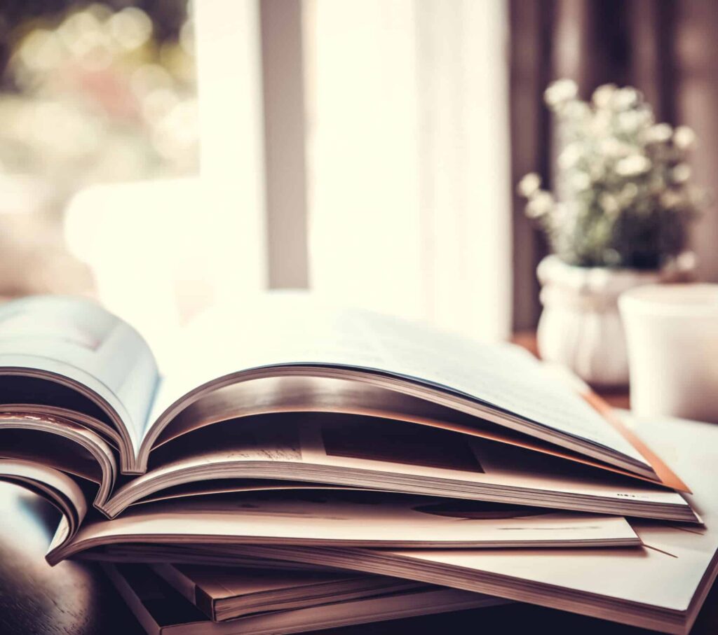 A close-up of an open book on top of a stack, with blurred indoor light and a small potted plant in the background, evoking a warm, cozy reading atmosphere—perfect for dreaming about recreational land or hunting property adventures.
