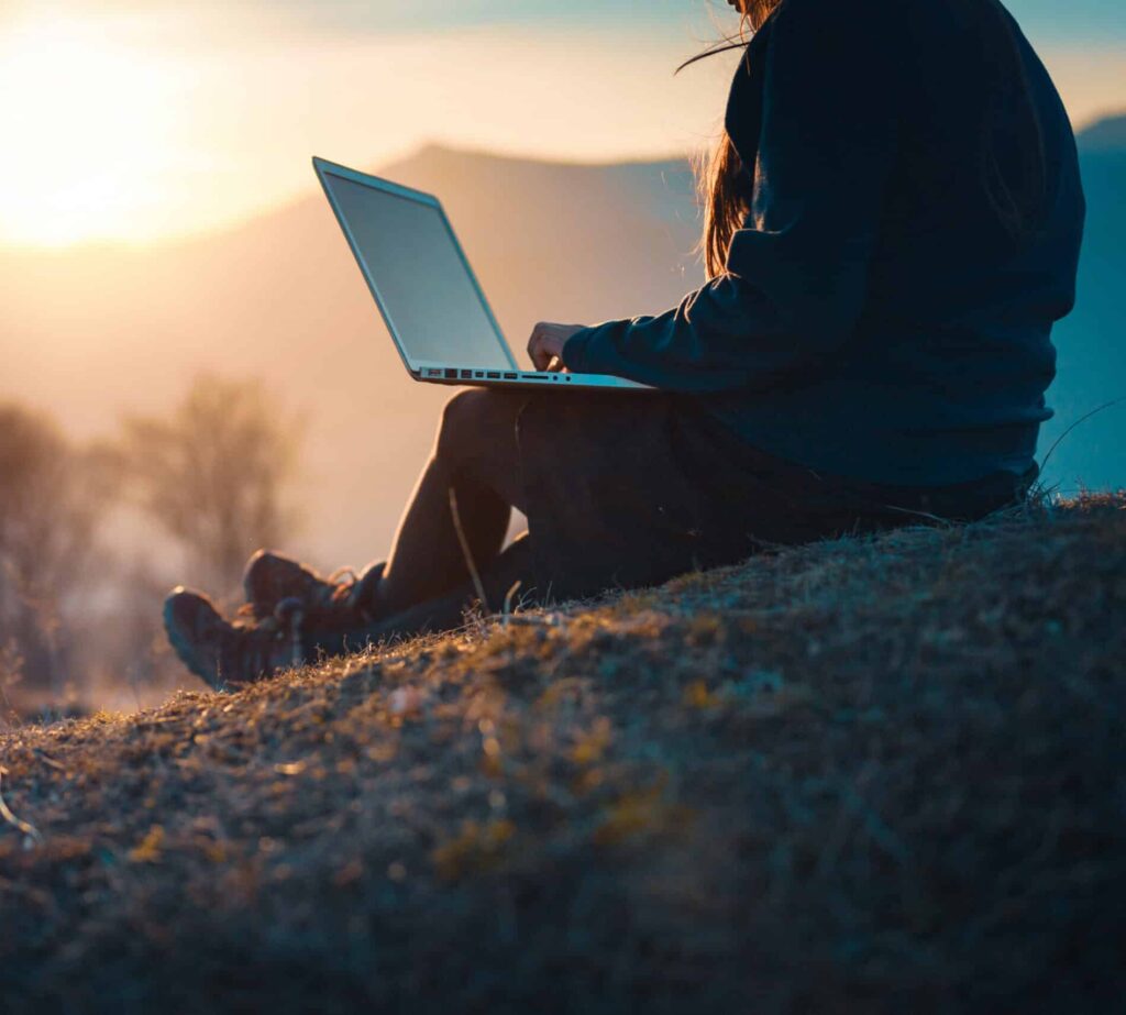 Person sitting on a grassy hill at sunset on recreational land, using a laptop. The scene is peaceful with soft sunlight, blurred trees, and distant mountains in the background. Only the person's silhouette is visible.