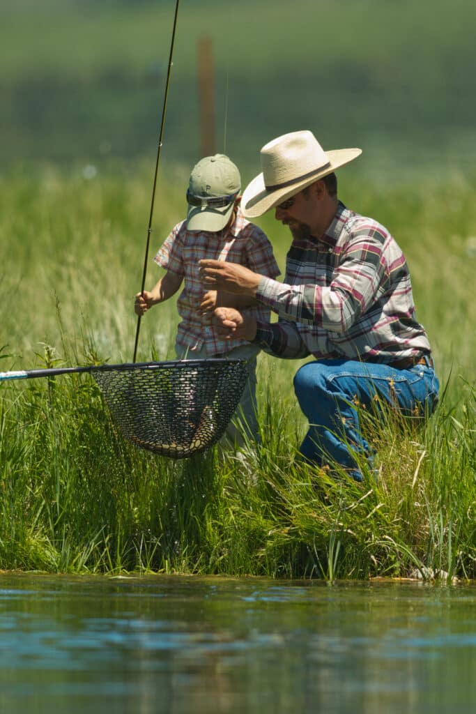 A man in a cowboy hat and a young child stand by a riverbank on recreational land, fishing together. The man helps the child with a fishing rod as a fish rests in a net above the water. Both wear plaid shirts and caps.