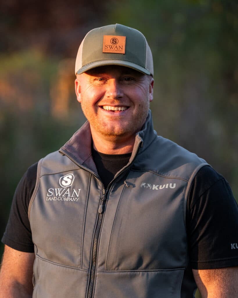 A smiling man wearing a gray cap and a gray sleeveless vest with Swan Land Company and KUIU logos stands outdoors in natural sunlight, representing recreational land and hunting property.