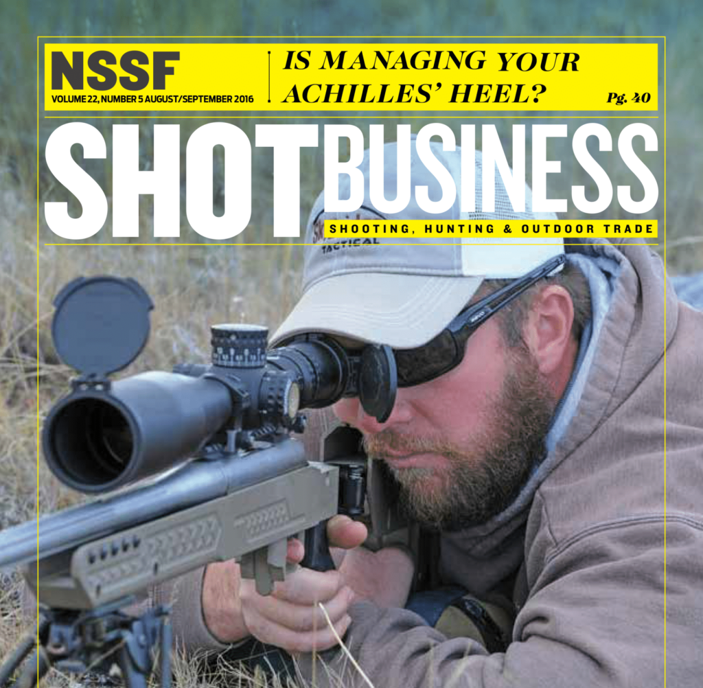 A man in tactical gear aims through the scope of a sniper rifle while lying prone on recreational land. The cover features the magazine title SHOT BUSINESS and other text.