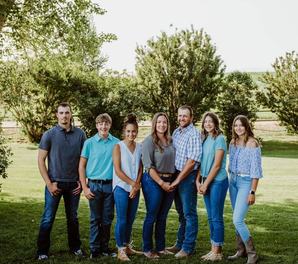 Seven people stand side by side outdoors on grass, with trees and fields in the background—ideal recreational land. They are casually dressed in blue jeans and various tops, smiling and facing the camera.
