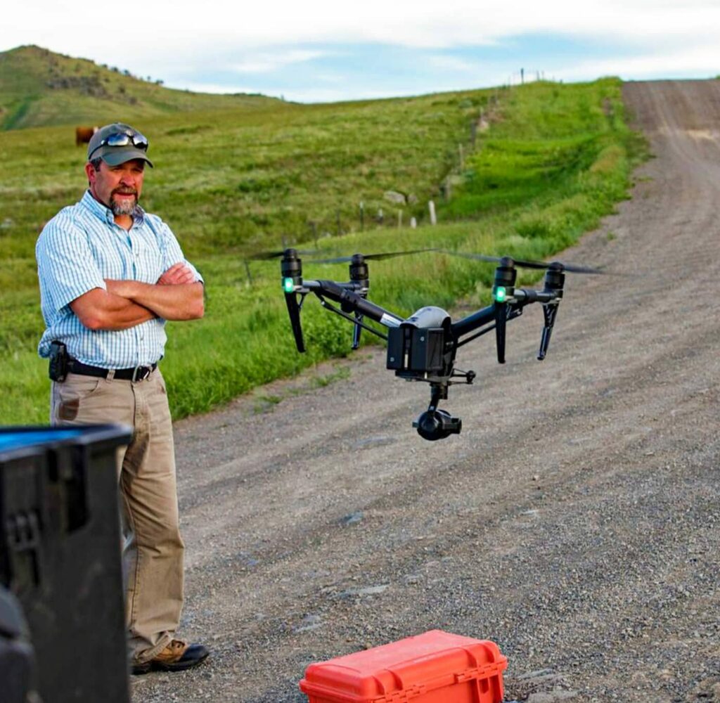 A man stands on a dirt road with arms crossed, watching a black drone hover in front of him. A red case rests on the ground, with grassy hills in the background—an ideal setting for exploring recreational land under a partly cloudy sky.