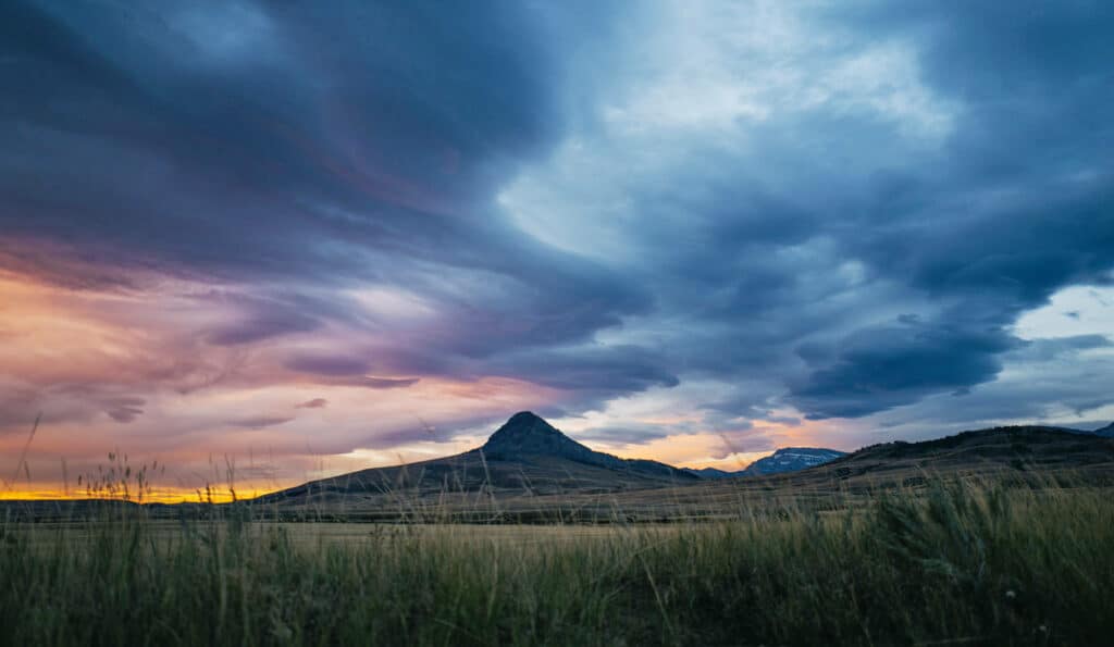 A dramatic sunset sky with vibrant blue, purple, and orange clouds stretches over a grassy plain on a scenic cattle ranch, with a pointed mountain peak rising in the distance.