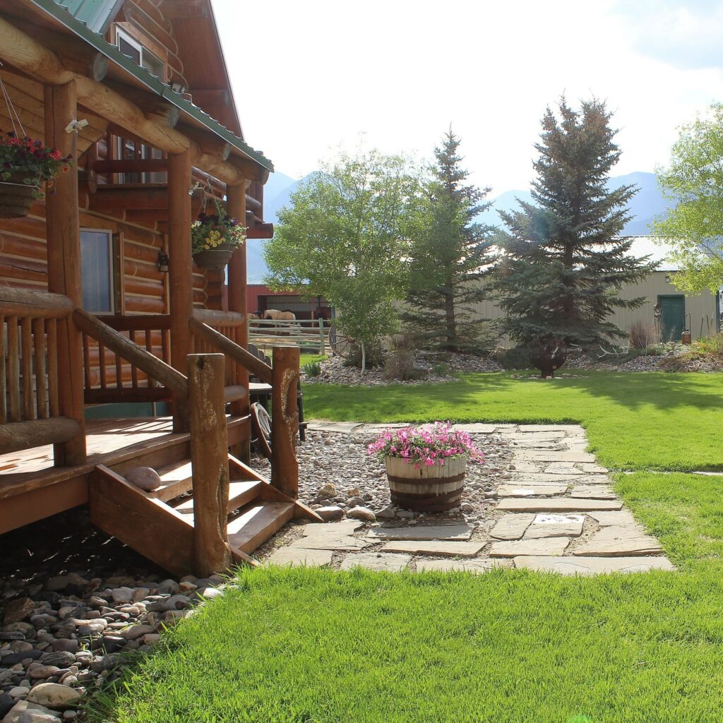A rustic log cabin with a wooden porch overlooks a green lawn, stone pathway, and trees. Perfect as a ranch for sale, it features a wooden barrel planter with pink flowers near the steps while sunlight brightens the scene.