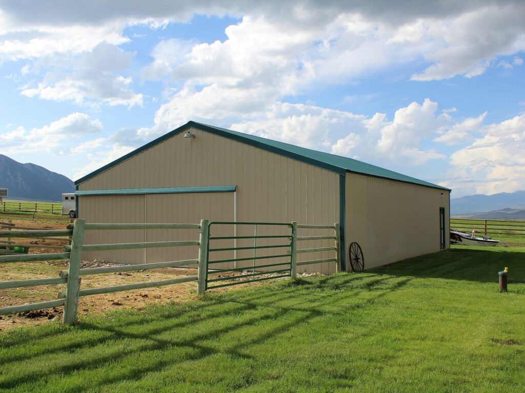 A large beige metal barn with a green roof stands on a grassy lawn, surrounded by a wooden fence. Mountains and a partly cloudy sky complete this scenic ranch for sale.