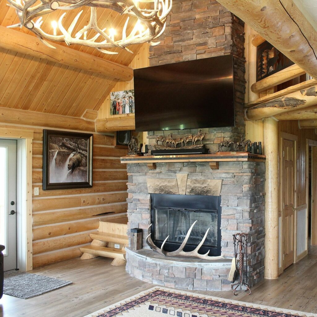 A cozy log cabin living room featuring a stone fireplace with antlers, a mounted TV above, wooden beams, animal-themed décor, and warm natural wood walls—perfect for relaxing after exploring nearby hunting property or land for sale.
