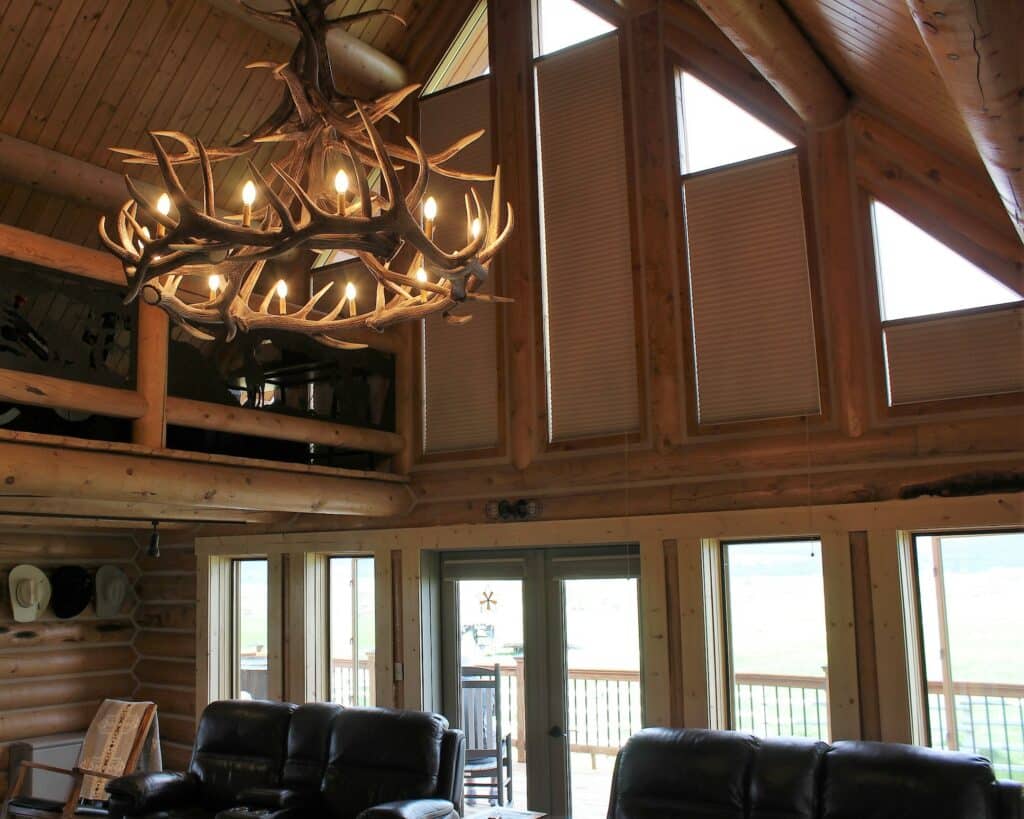 A cozy log cabin interior with high, slanted wooden ceilings and leather sofas sits on recreational land. Large windows with closed blinds and glass doors bring in natural light, while an antler chandelier hangs from above.