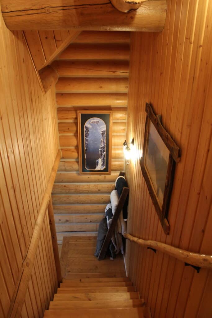 A wooden staircase in a log cabin with rustic log walls, a framed picture of a snowy mountain scene at the bottom, wall-mounted light, and several rolled-up blankets in the corner—perfect inspiration for your own recreational land retreat.