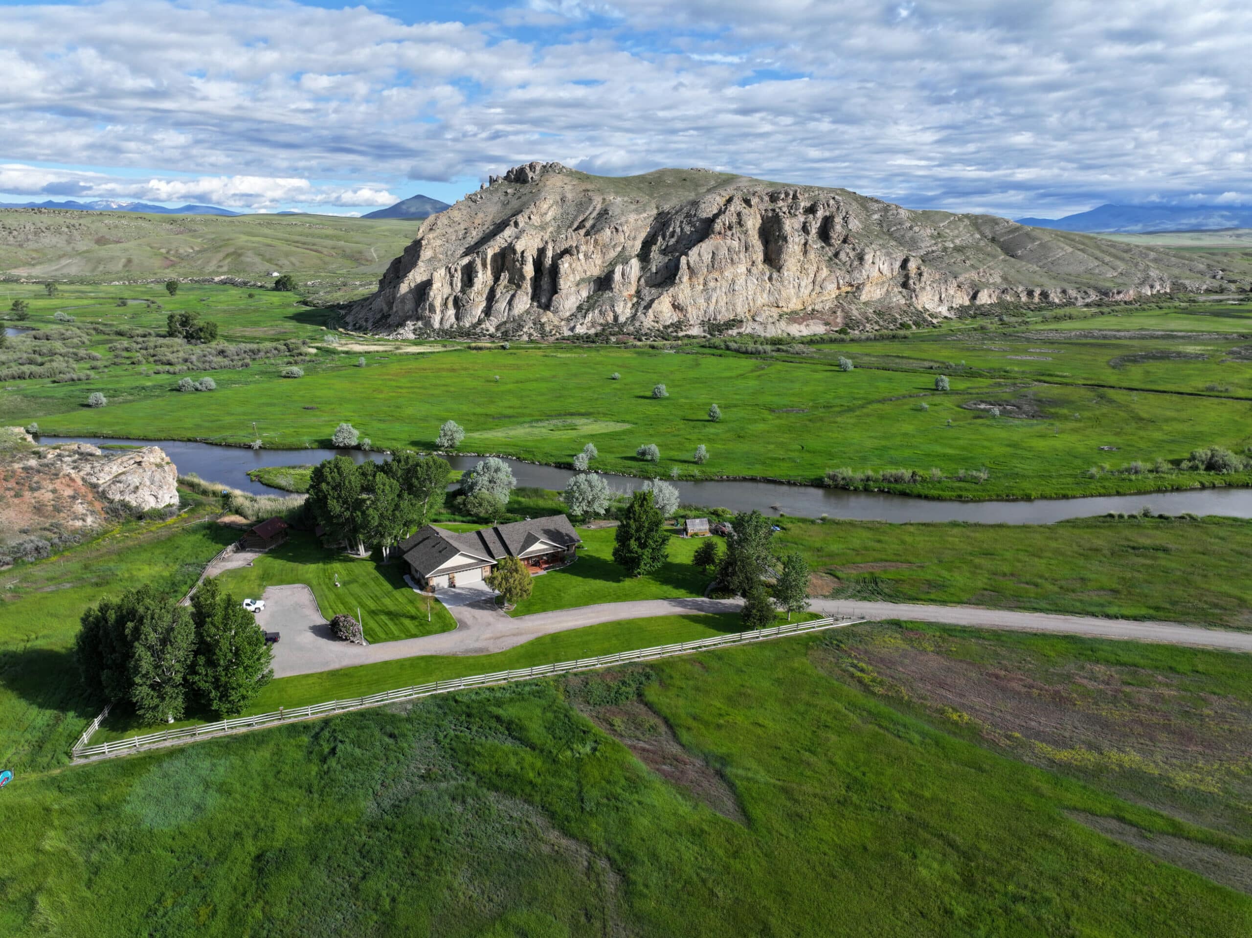 Aerial view of a ranch for sale surrounded by green fields, trees, and a winding river, with a large rocky hill and mountains in the background under a partly cloudy sky.