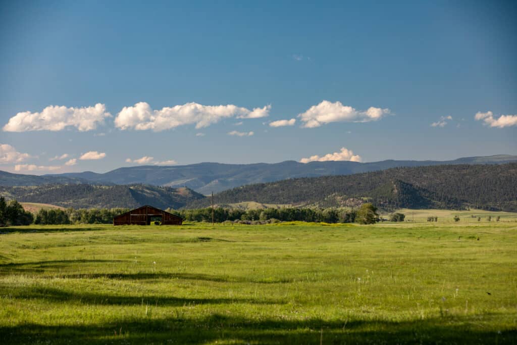 A red barn sits in a wide green field under a blue sky with scattered clouds, surrounded by forested hills and mountains—perfect as a cattle ranch or hunting property, with land for sale in the scenic countryside.