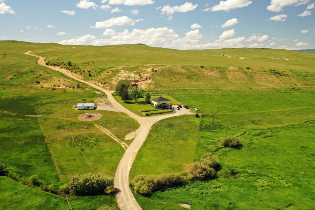 Aerial view of a rural landscape with green fields, dirt roads, a small house surrounded by trees, and an outbuilding—perfect as a hunting property or ranch for sale—set beneath a bright, partly cloudy sky and gently rolling hills.