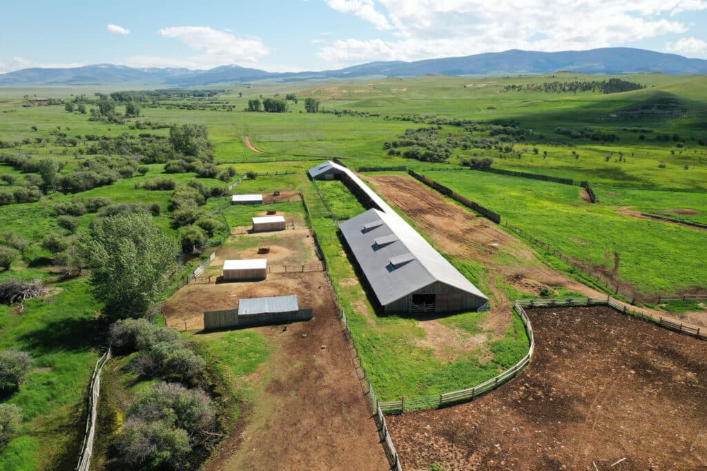 Aerial view of a rural farm with several barns, fenced areas, and open green fields surrounded by rolling hills and distant mountains under a partly cloudy sky—ideal ranch for sale or premium hunting property.