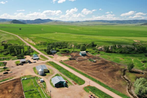 Aerial view of a rural ranch for sale with barns, outbuildings, fenced areas, dirt roads, and green fields stretching into the distance under a blue sky with scattered clouds.