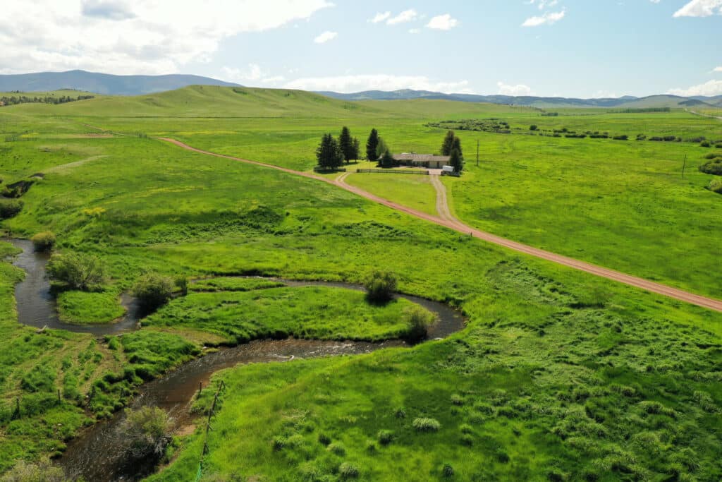 A small house surrounded by trees sits in the middle of vast green fields on recreational land, with a winding dirt road and a creek flowing through the grassy landscape under a partly cloudy sky.