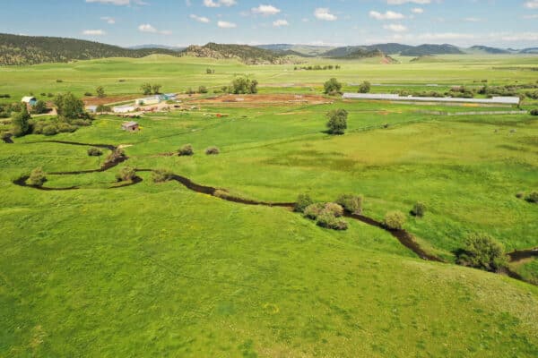 Aerial view of a green rural landscape with fields, a winding stream, scattered trees, and rolling hills in the background—ideal recreational land or cattle ranch under a blue sky with clouds.