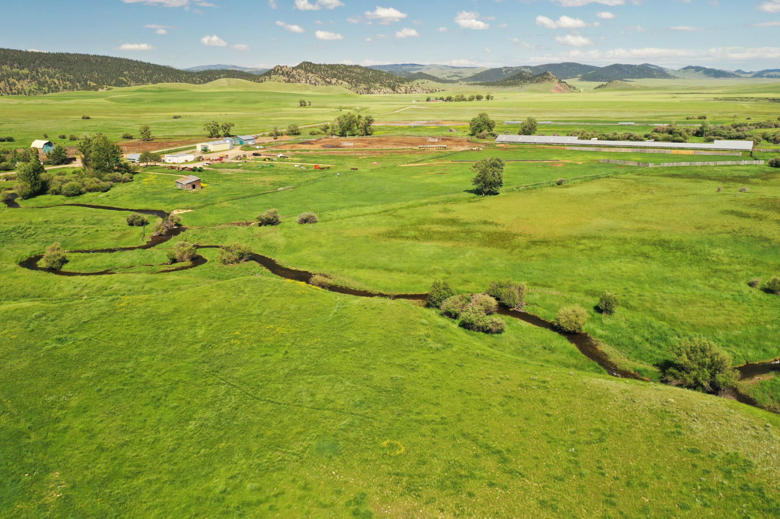 Aerial view of a green rural landscape with fields, a winding stream, scattered trees, and rolling hills in the background—ideal recreational land or cattle ranch under a blue sky with clouds.
