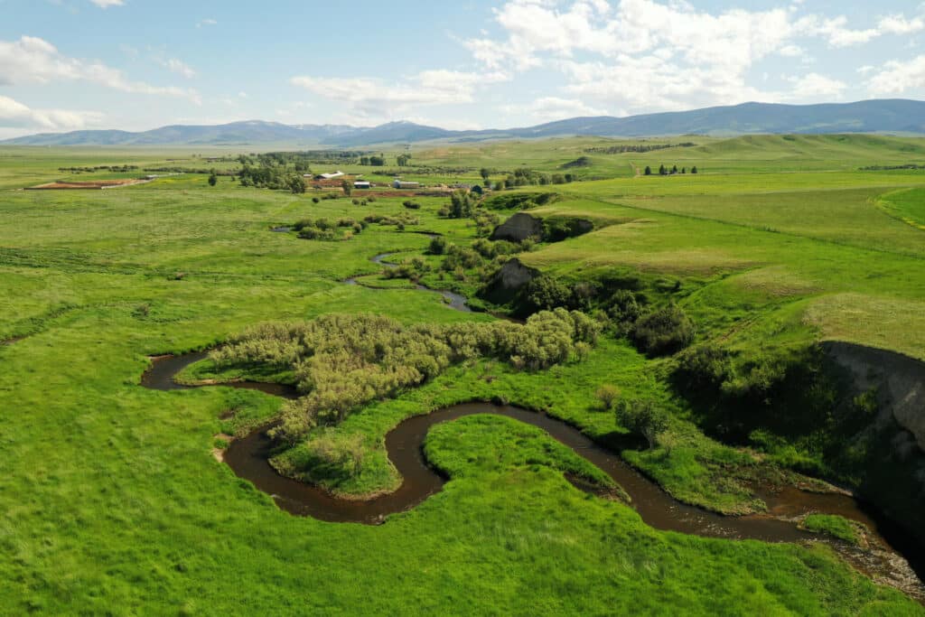 A winding stream flows through lush, green recreational land with grassy fields, scattered trees, and distant mountains under a partly cloudy sky. Farms and buildings are visible in the background.