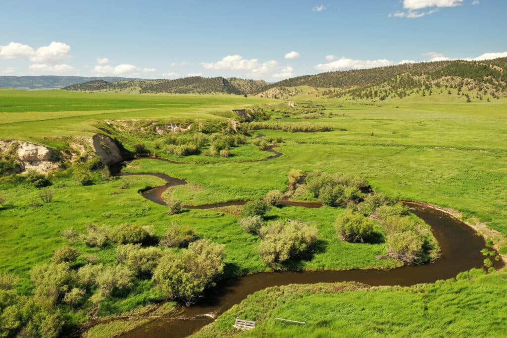A winding river flows through lush green meadows and gentle hills under a blue sky, surrounded by fields and distant tree-covered slopes—ideal recreational land or hunting property.