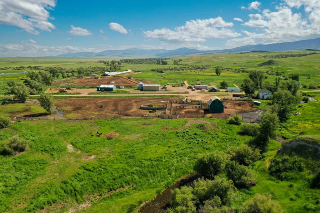 Aerial view of a rural ranch for sale with barns, fenced fields, green pastures, scattered trees, and distant hills under a partly cloudy blue sky.