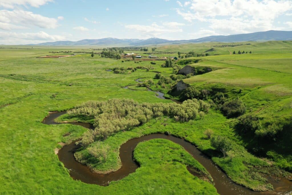 A winding stream flows through lush green fields with distant farm buildings and rolling hills, perfect for a cattle ranch, under a partly cloudy sky.