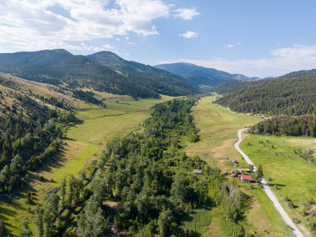 Aerial view of a green valley bordered by forested mountains, featuring recreational land with a winding road leading to a small cluster of red-roofed buildings under a partly cloudy sky.