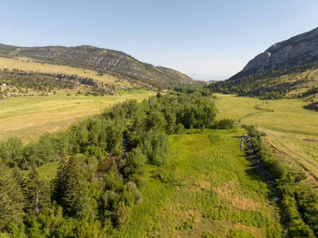 Aerial view of lush, green recreational land with a stream winding through grassy meadows and dense trees, surrounded by hills and rocky cliffs under a clear blue sky.