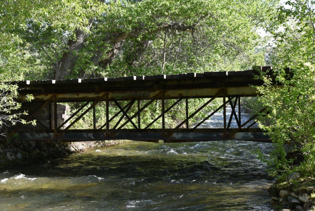 A metal and wood bridge crosses a flowing river, surrounded by lush green trees on land for sale, with dense foliage stretching beneath the sunny sky.