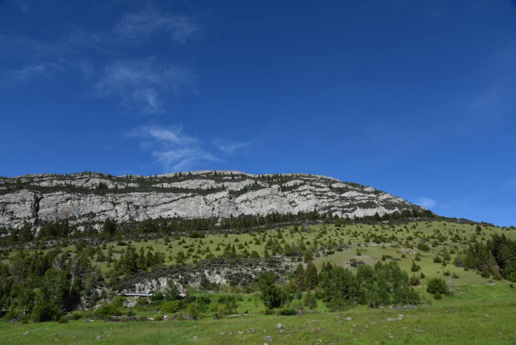 Rolling hillside with scattered green trees leads up to a large, rocky, layered mountain under a vibrant blue sky—ideal hunting property or land for sale for those seeking natural beauty and adventure.