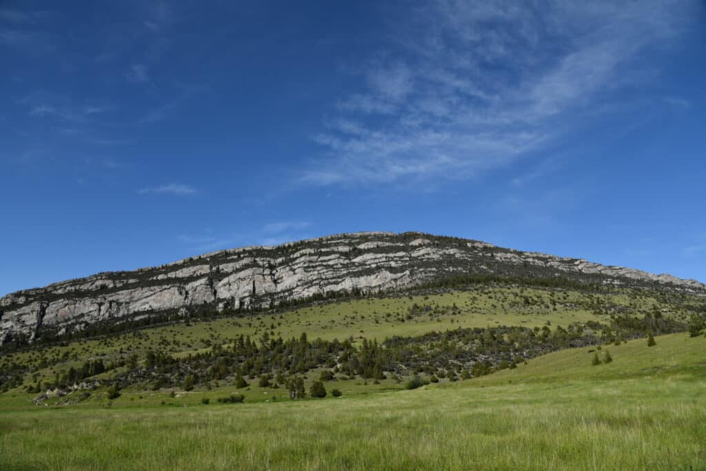 A grassy field leads up to a hill with exposed, layered rock formations and scattered trees under a bright blue sky—ideal recreational land or hunting property.