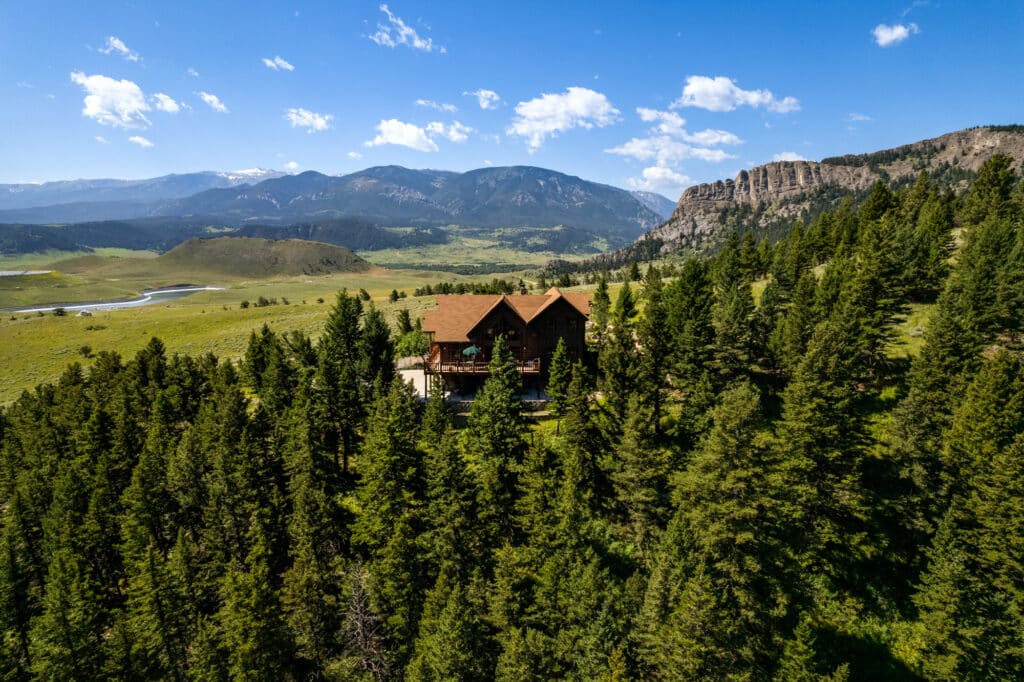 A large wooden cabin surrounded by dense evergreen trees sits in a lush valley, with mountains and rocky cliffs in the distance under a blue sky—perfect for those seeking a scenic cattle ranch or unique land for sale.