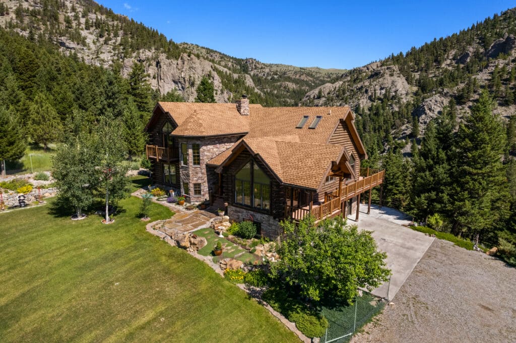 A large log cabin-style house with stone and wood exterior sits amid green grass and trees on recreational land, surrounded by rocky mountains under a clear blue sky. A driveway and landscaped yard are visible.