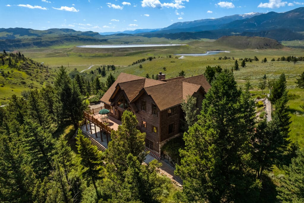 A large wooden cabin surrounded by tall trees sits on a hillside overlooking a vast green valley, perfect as recreational land or hunting property, with mountains and a lake visible in the background under a blue sky with scattered clouds.