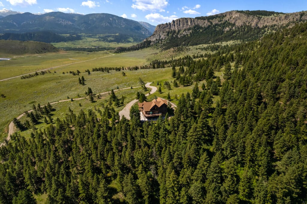 A large rustic lodge is nestled among dense evergreen trees in a wide valley, surrounded by mountains and open grassy fields—an ideal setting for a cattle ranch under a partly cloudy sky.