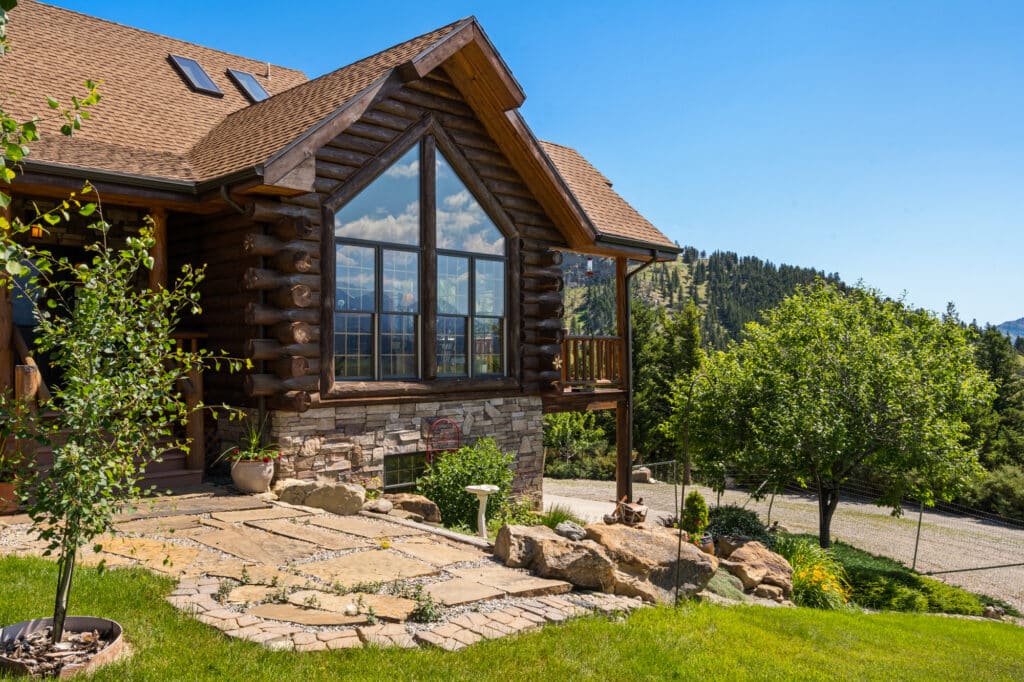 A rustic log cabin with large windows, stone foundation, and manicured garden sits on a grassy hillside of a serene cattle ranch for sale, overlooking distant forested mountains under a clear blue sky.